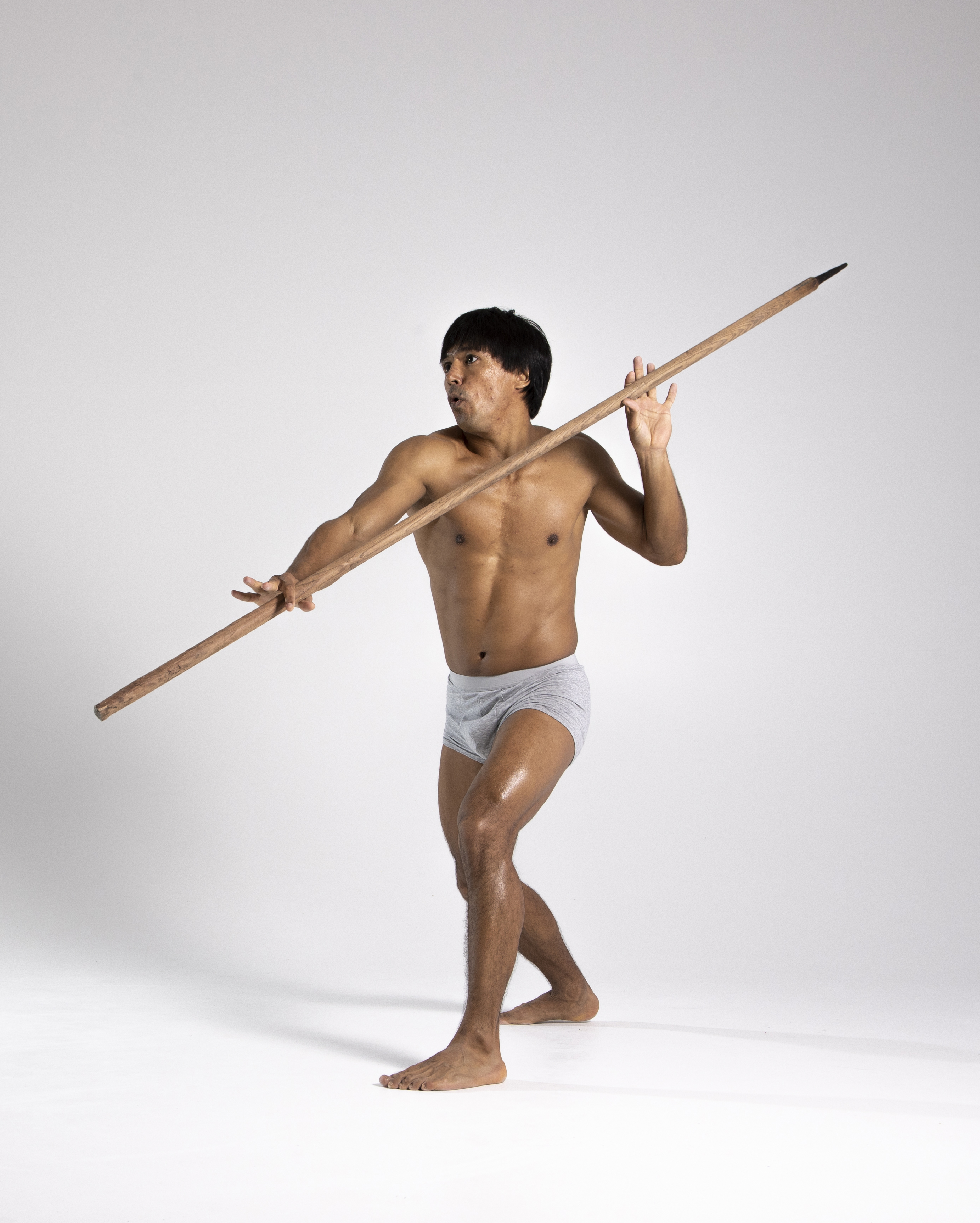 a shirtless man wearing a loincloth is holding a spear in a studio setting against a white background. he appears to be an indigenous person and is in a hunting pose.