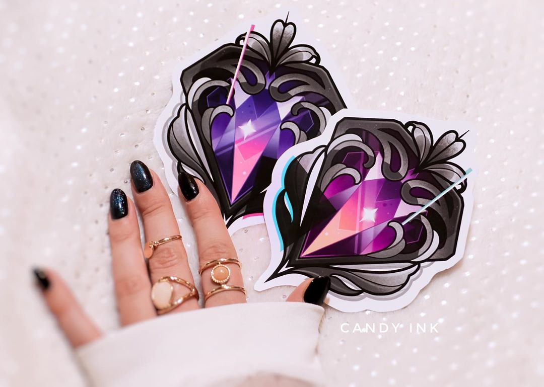 a woman's hand with rings and dark nail polish is holding two identical stickers against a white, textured background. the stickers feature a stylized design of a pink and purple gemstone set in an elaborate, black and grey,  vintageinspired frame. the stickers are from a brand called "candy ink".