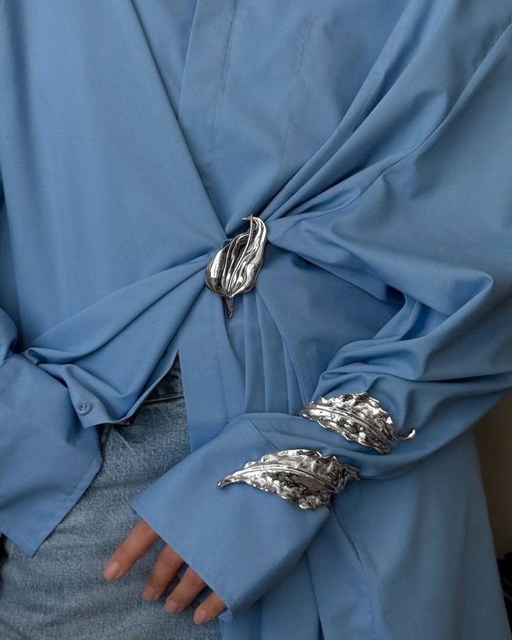 closeup shot of a light blue shirt with silver leafshaped jewelry. one leaf brooch is used to fasten the center of the shirt, and two leaf cuffs are used to fasten the sleeves. the person wearing the shirt is also wearing denim pants.