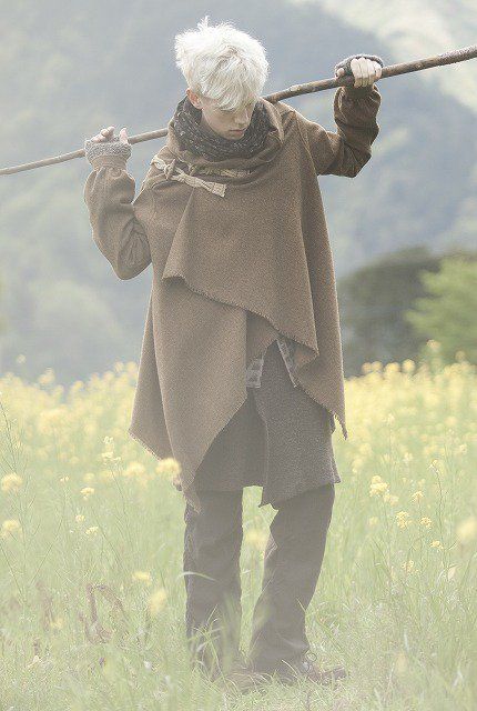 a man with white hair is walking through a field carrying a staff. he is wearing a brown shawl and scarf. the landscape is overcast.