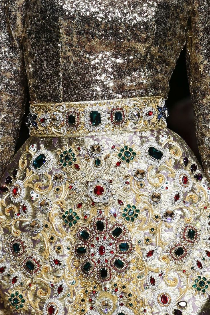 loseup shot of a lavish, intricately detailed gown. the dress is predominantly gold, covered in sequins and sparkling embellishments. a wide, ornate belt is adorned with large gemstones and intricate gold work. the skirt of the dress features detailed embroidery and a profusion of jewels, including emeralds, rubies, and clear crystals, creating a rich, opulent look. the overall texture is luxurious and shimmery, conveying a sense of high fashion and elegance.