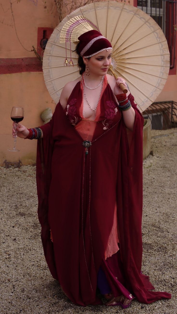 a woman in a long, flowing burgundy dress and a matching head covering stands outdoors, holding a parasol. she is wearing various jewelry pieces and holds a glass of red wine. the setting appears to be historical or fantasythemed. the dress is elegant and detailed with embroidery. the overall impression is of a stylish and mystical figure.