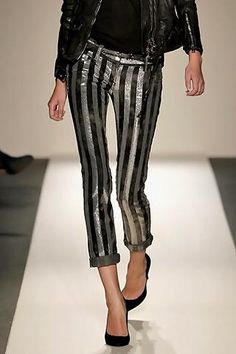 a woman in a black leather jacket and black and white striped pants walks on a runway during a fashion show. she is wearing high heels. she's posing for the cameras.  the woman's style is chic and trendy.
