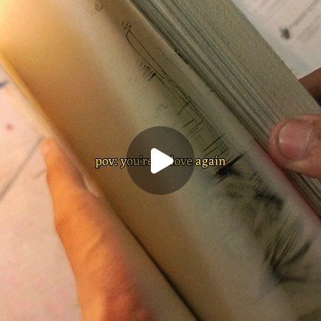 the image shows a closeup of a person's hands interacting with a wellworn, possibly antique book. the pages are aged and show signs of use, including markings and scribbles. a play button is overlaid on the image, along with the text "pov: you're in love again". the overall mood is nostalgic and romantic, suggesting a feeling of rediscovering love or a past relationship. the setting appears informal, possibly a personal video or social media post.
