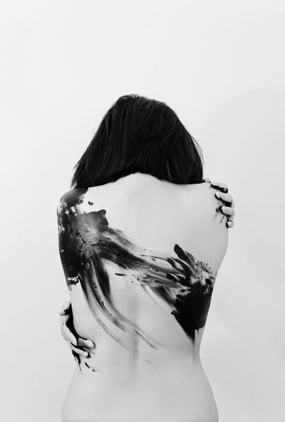 a black and white portrait of a young woman standing in profile against a white background. the woman's back is covered in black paint that creates an abstract design.  the  woman's hair is long and black.  she is looking at the camera.  the image is taken in a studio setting.