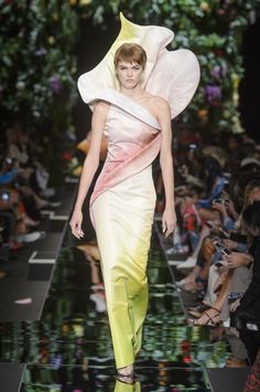a fashion model walks the runway in a white, yellow, and pink floral dress. the dress is made from a single large flower and is very unique and avantgarde. the model is wearing her hair straight and is looking down at the runway. the background is blurry and shows other people watching the show.