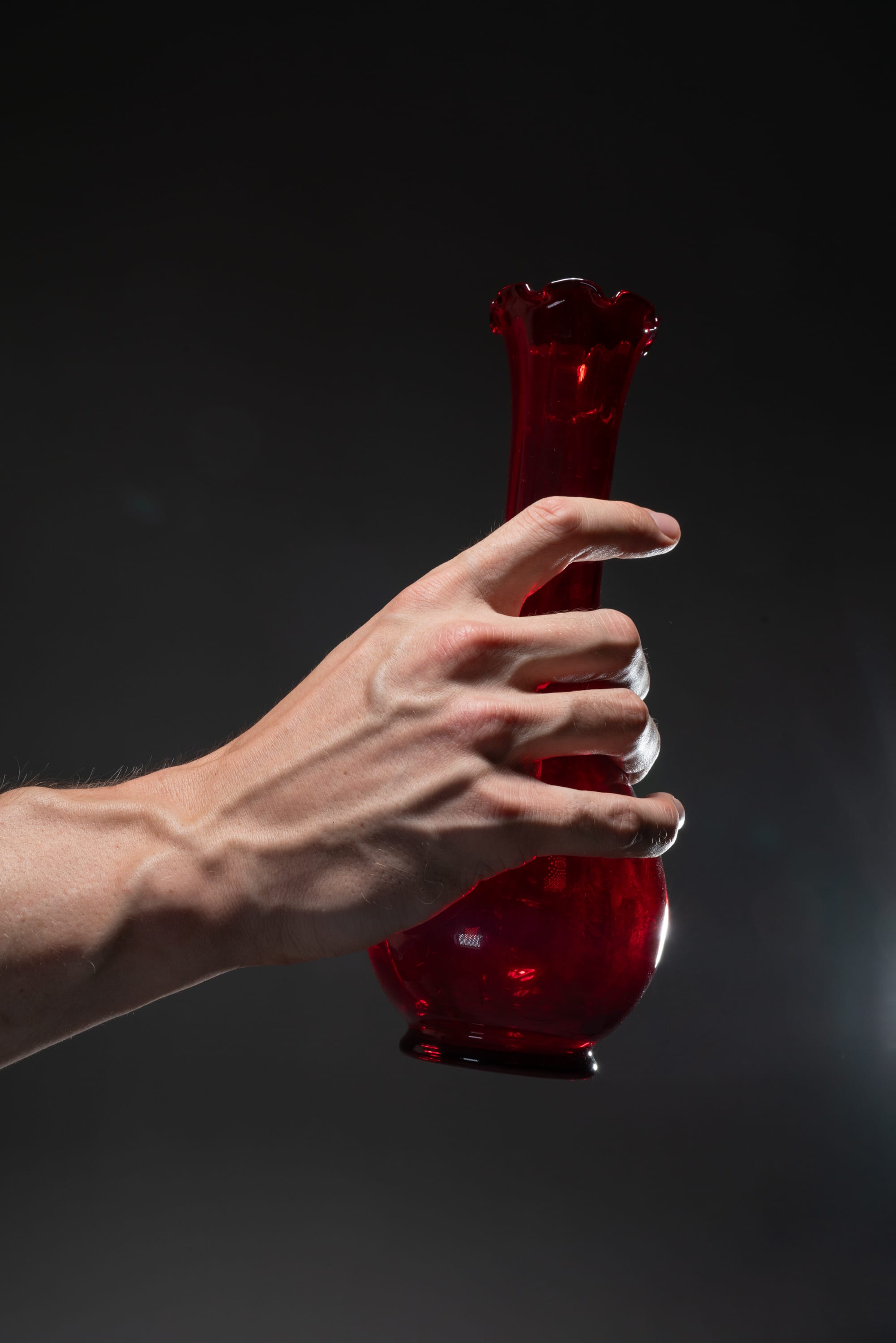 a caucasian male hand holding a red glass vase against a dark gray background. the image is lit with studio lighting, highlighting the vase and the veins in the hand.