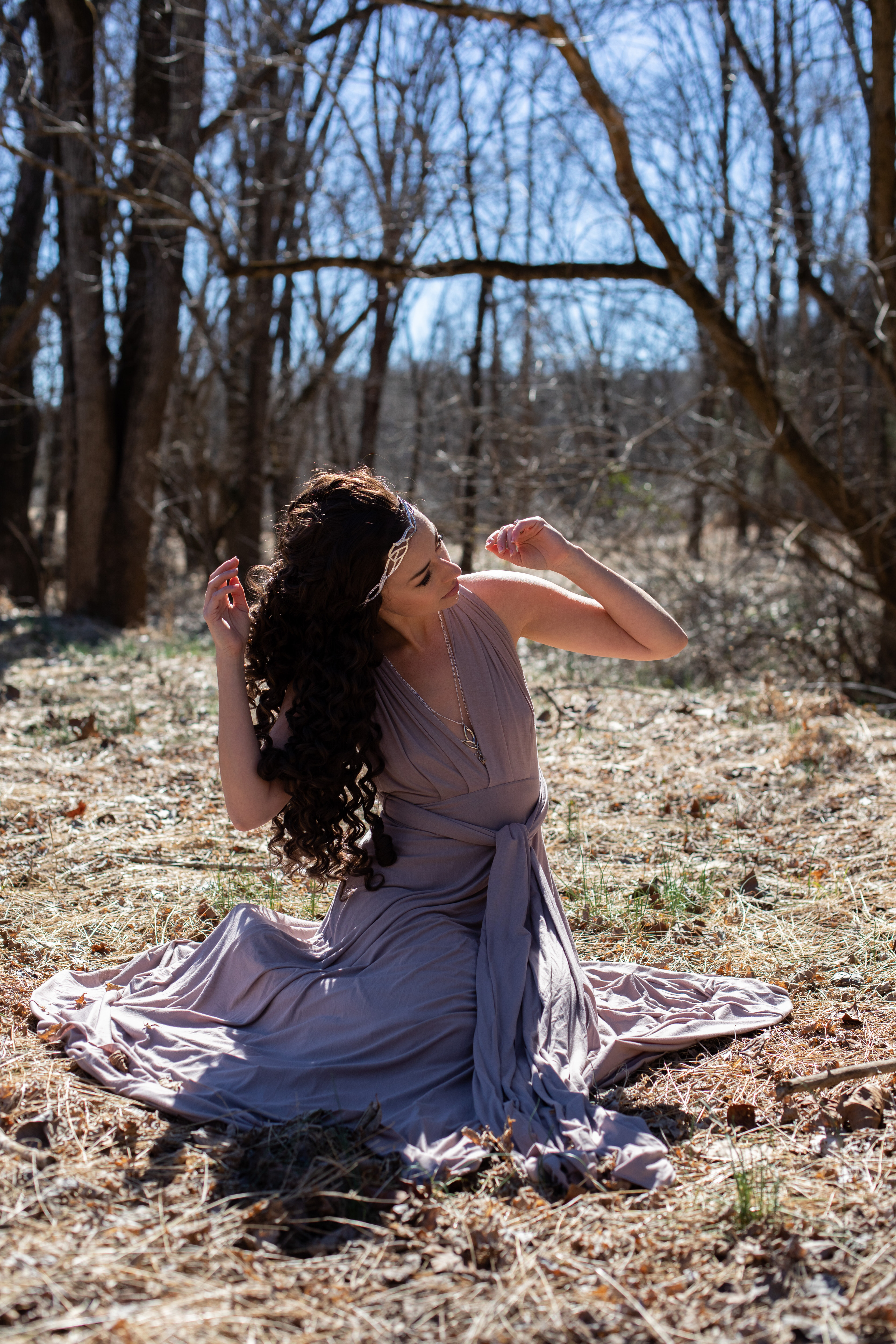 a young woman with long, dark curly hair, adorned with a delicate headpiece, sits gracefully on the forest floor. she wears a flowing, lavendercolored gown that spreads out around her, complementing the natural beauty of the surrounding woods. the sunlight peeks through the trees, casting warm shadows on the ground and illuminating her features. her pose is elegant and contemplative, suggesting a connection with the tranquility of nature.