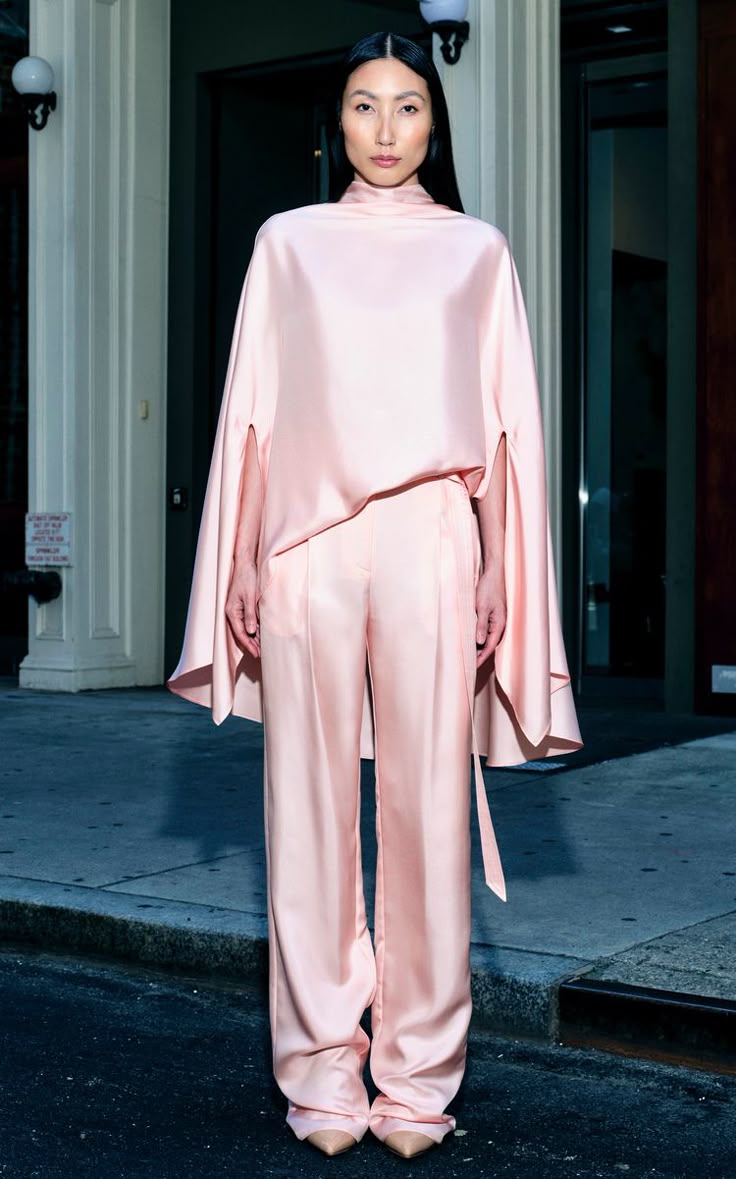 a full shot features a woman posing in a pink satin fashion ensemble consisting of a cape, trousers, and shoes. she is standing on a street with buildings in the background. the woman has dark hair and is looking directly at the camera. her pose is straighton, with her arms relaxed at her sides. the cape drapes over her shoulders, and the trousers are widelegged. the shoes are pointytoed and appear to be the same material and color as the rest of the outfit. the street is slightly darker than the surrounding buildings, and the architecture is a mix of traditional and modern. the lighting is soft and even, highlighting the shine of the satin material. the overall impression is one of sophistication and style.