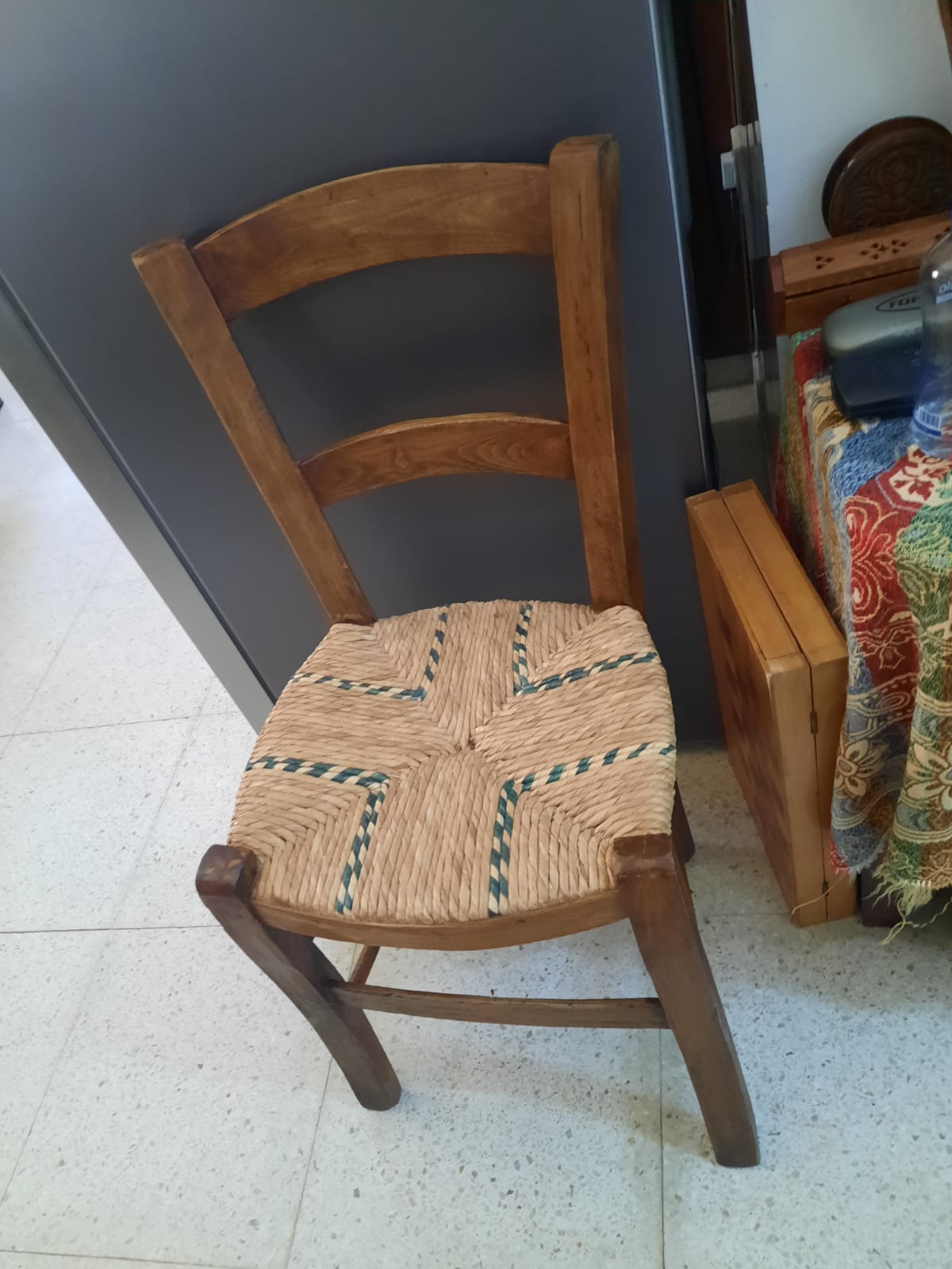 a wooden chair with a woven straw seat is positioned on a tiled floor, leaning against a grey cabinet. a colorful cloth and a wooden box are visible beside the chair.