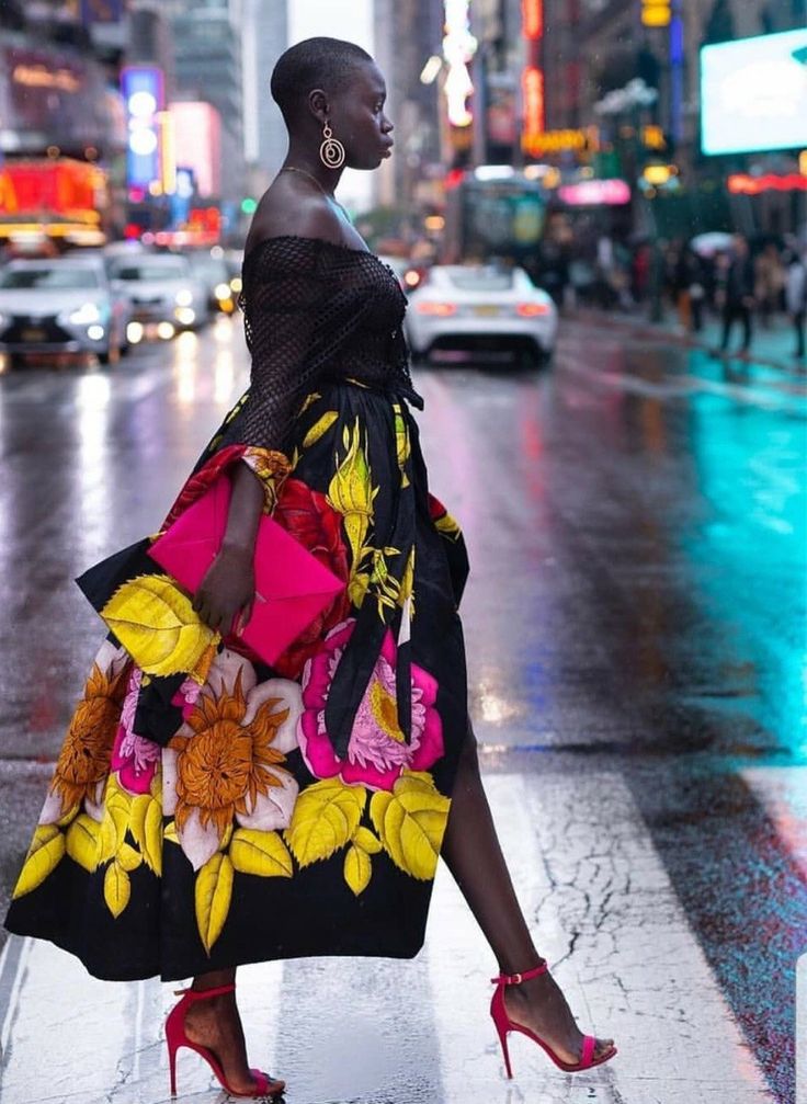 a stylish woman in a black floral dress with a pink clutch and red heels walks confidently across a street crossing in a city setting. she has short hair and is wearing earrings. the scene is urban and the background is blurred. the woman is the focal point of the image.
