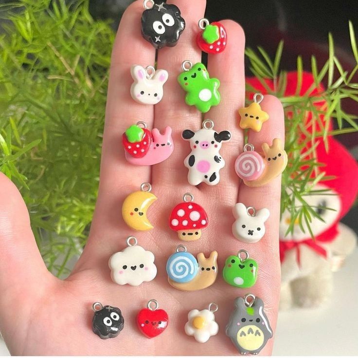 a hand holding a collection of handmade polymer clay charms in various cute and kawaii designs including a strawberry, snail, cow, bunny, mushroom, totoro, star, moon, cloud, flower, heart, and frog.