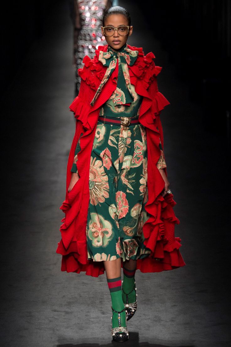 a runway model walks down the catwalk in a striking gucci outfit.  the ensemble features a long green floralprint dress under a voluminous red ruffled cape. she accessorizes with green and red striped socks, jeweled heeled shoes, and goldframed glasses. a guccibranded belt is visible at her waist. the overall style is a blend of vintage and modern, with a bohemian flair.  the background is dark, focusing attention on the model and her outfit.