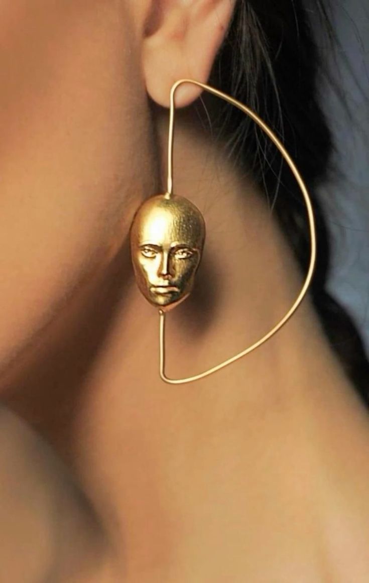a woman is wearing a gold earring with a face pendant and a geometric design. the earring is a contemporary style with a minimalist design. the woman's skin is fair and the earring is a statement piece.