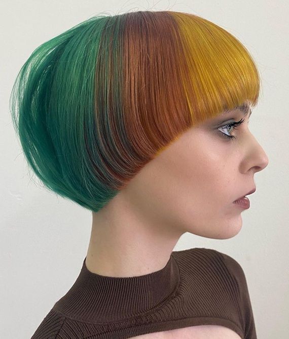 a young woman with short, colorful hair. her hair is cut in a bob style with blunt bangs. the hair is dyed in shades of green, yellow, orange and brown. the woman is wearing a brown turtleneck sweater and has natural makeup on.
