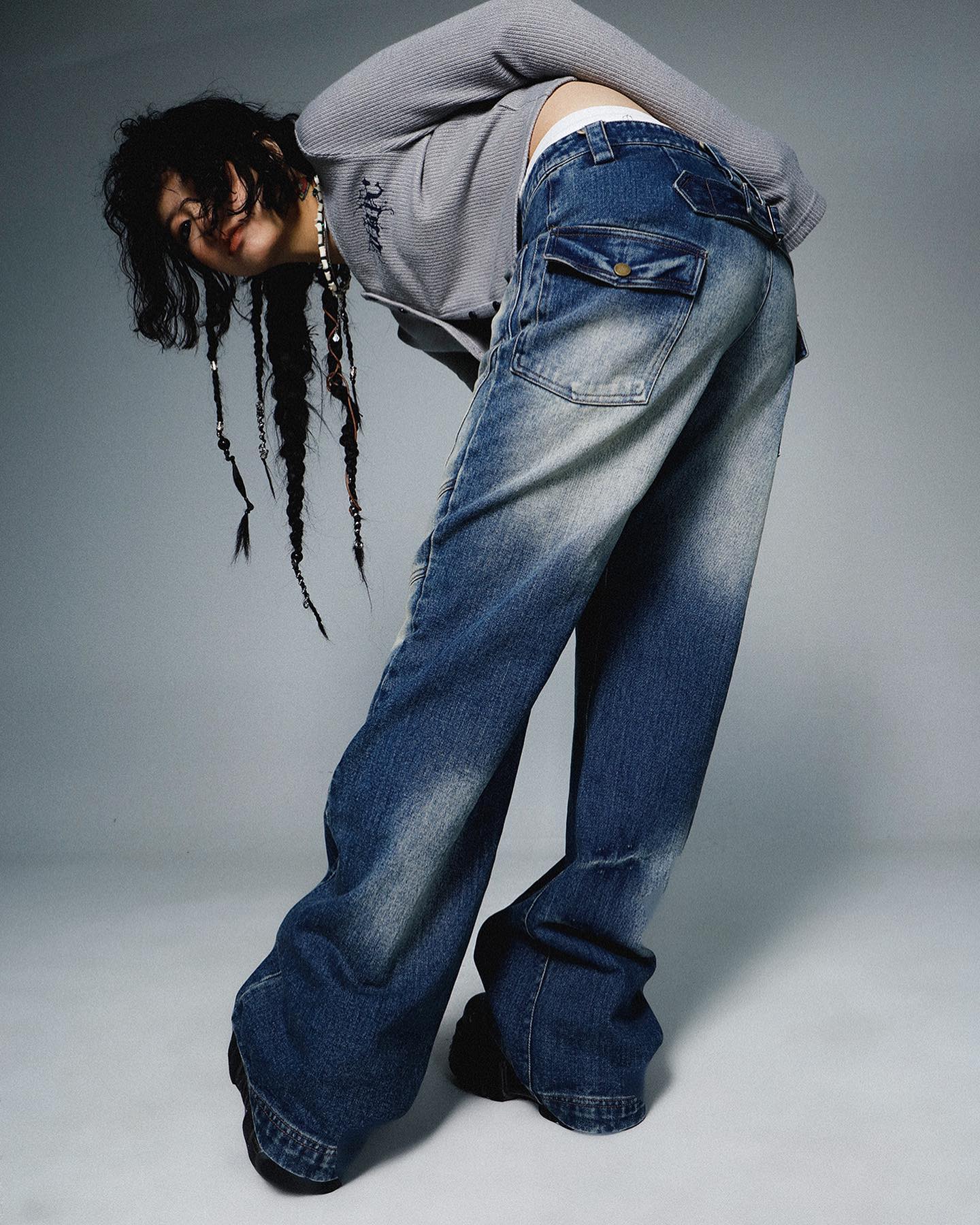 a fashion photograph showcases a young woman with long braided hair, wearing a loosefitting gray longsleeved sweater and a pair of baggy, wideleg, distressed blue denim jeans. the jeans have prominent pockets and a low waistline. the model is posing in a studio setting with a gray backdrop, and her pose leans forward, showcasing the drape and texture of the jeans. the overall style is casual and trendy, with a hint of vintage feel.  the image appears to be professionally styled for a fashion editorial or catalog.