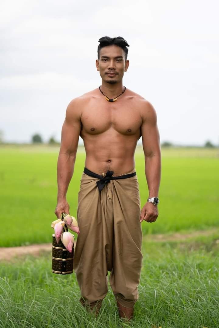 a man stands in a rice field. he is shirtless, showing off his muscular build. he is wearing brown pants and is holding a basket of pink lotus flowers. the man is smiling and looking directly at the camera. his arms are at his side. the background is a lush green rice field with a blue sky. he appears to be very confident and happy.