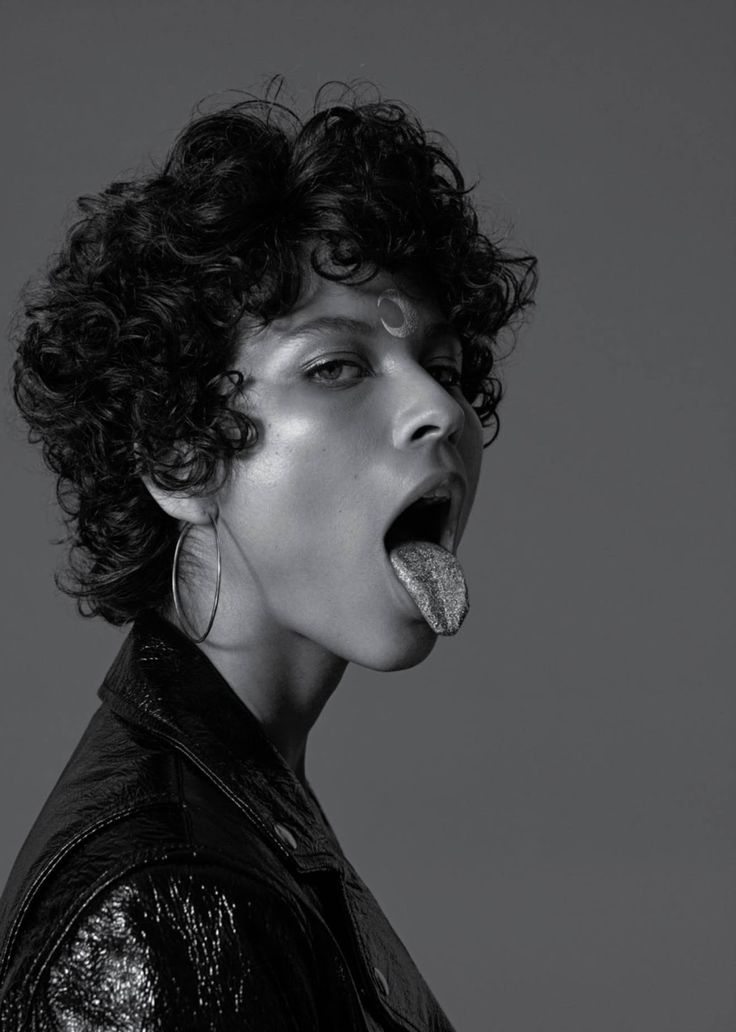 a black and white portrait of a woman in a leather jacket sticking out her tongue and looking away. she is wearing makeup, eyeshadow and lipstick, and has short curly hair with a large earring on her left ear. she is standing in a studio against a grey background.