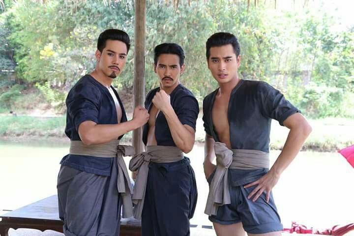 three young men, dressed in traditional thai clothing,  stand together,  smiling at the camera,  outdoors in natural light.   the men are all asian, wearing black and blue clothing,  with a brown sash around their waist. the men appear to be friends and are posing for a photo.