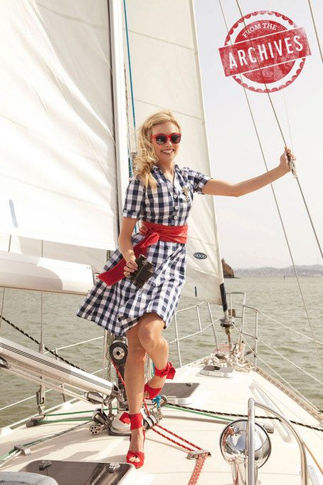 a woman in a gingham dress and red sandals is standing on the deck of a sailboat. she is holding onto a rope and smiling at the camera. she is wearing red sunglasses and has a red bow tied around her waist. the sailboat is docked in a harbor. the water is blue and the sky is sunny. the woman is enjoying a day of sailing on the water.