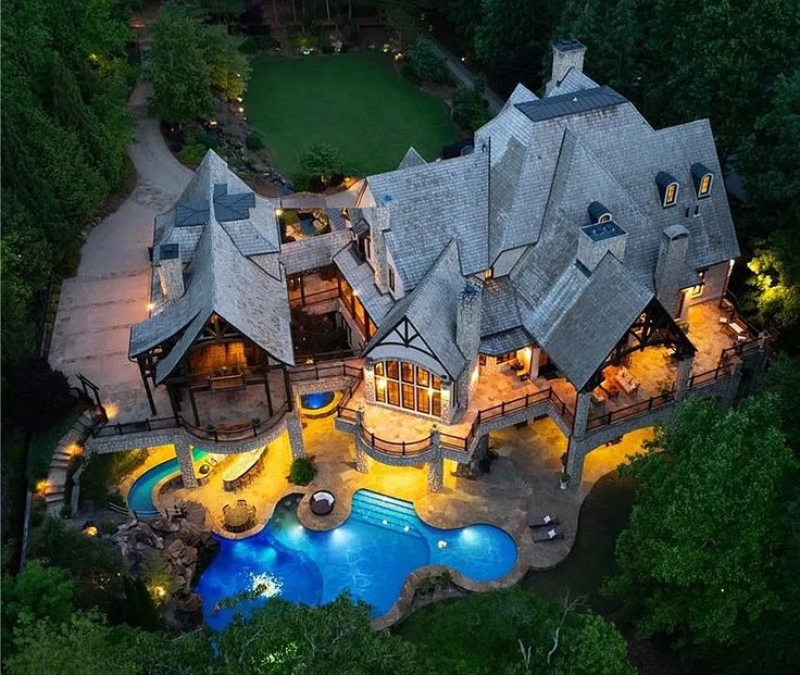 aerial view of a large, custombuilt luxury mansion at night with extensive outdoor lighting. it features a swimming pool, stone patio, slate roof, multiple chimneys, dormer windows, covered porches, decks, and detailed landscaping. the house is surrounded by trees and a wellmanicured lawn.