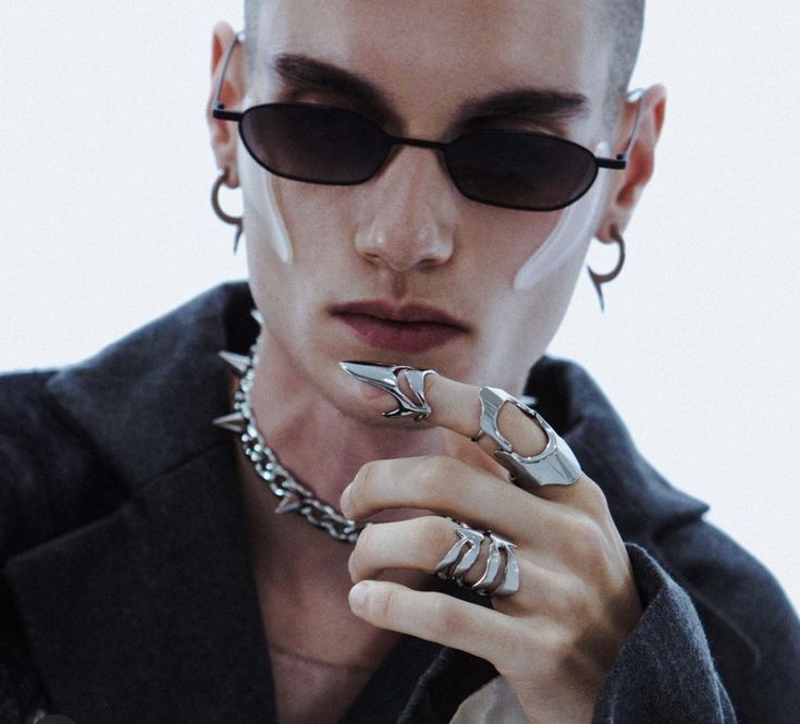 a young man is posing for a portrait while wearing a pair of black sunglasses, a variety of silver rings, and a black jacket. the man's style is trendy, edgy and cool. his expression is serious and brooding as he looks away from the camera. he is dressed in an alternative style that is both stylish and unique.