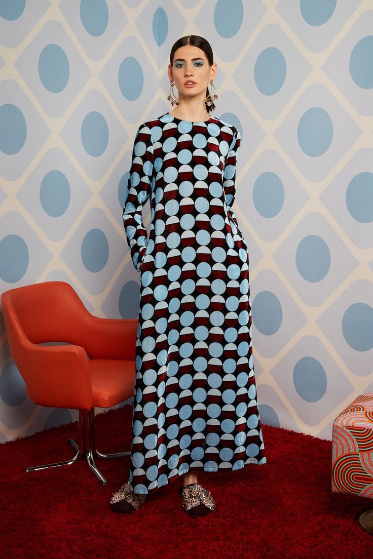 a woman is standing in a maxi dress with a geometric pattern of blue and maroon circles on a velvet fabric. she is wearing sparkly shoes and long earrings. she is posing in a studio setting with a colorful background, including a red chair and an ottoman, and a red carpet. the style is reminiscent of retro fashion from the 60s and 70s. the image is professionally shot and shows the dress and accessories in detail.