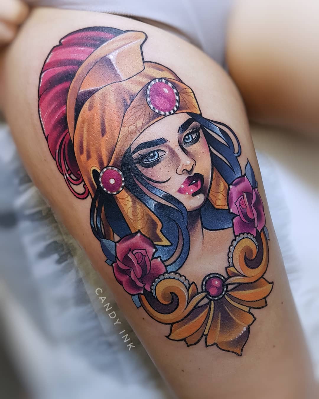 a colorful neotraditional tattoo of a woman's portrait on someone's upper thigh. the woman has dark hair and blue eyes and wears a gold greekstyle helmet. she is surrounded by roses and ornate gold jewelry with pink jewels.  the tattoo is credited to "candy ink."