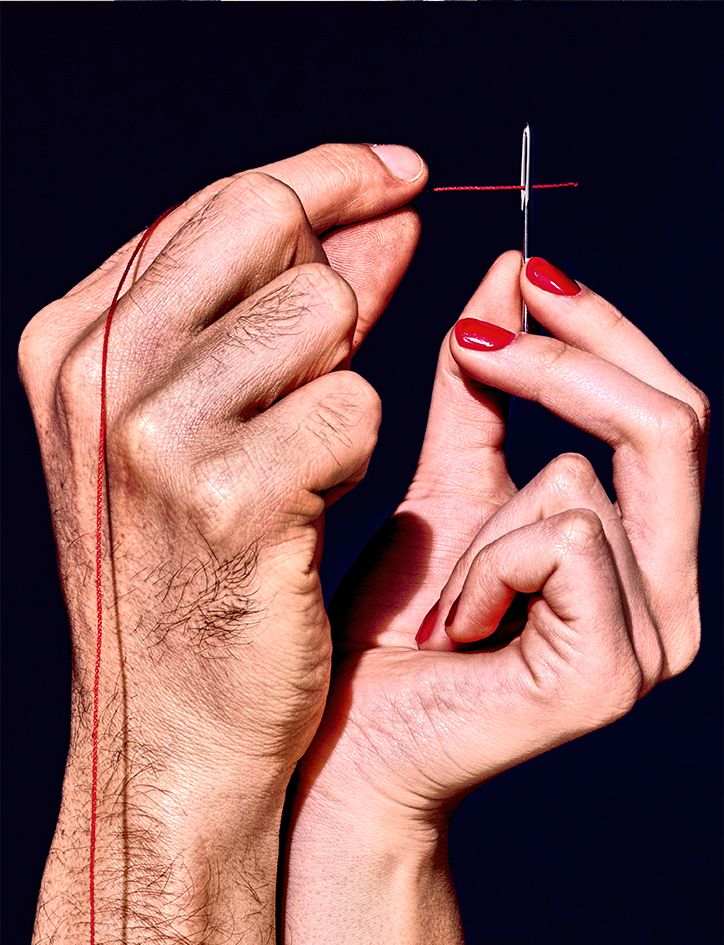 a closeup image of two hands, one with red nail polish and the other with no nail polish, intertwined. a red thread is being threaded through a needle, held in the hand with red nail polish. the hands are positioned in a way that symbolizes connection and unity.