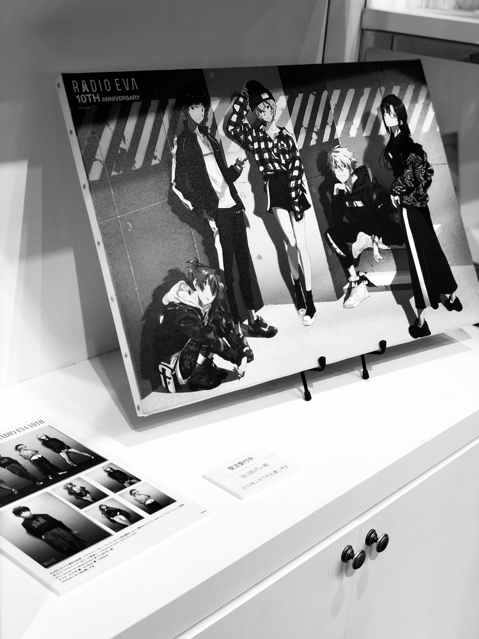 the image is a black and white photograph of a display case featuring a poster commemorating the 10th anniversary of radio eva. the poster, done in a manga style, depicts four characters posing against a striped backdrop. below the poster is a smaller print showcasing individual character shots. the display case itself is white and welllit, suggesting an indoor environment like a store or gallery.
