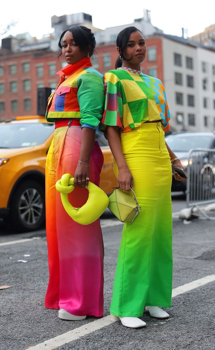 two women stand sidebyside on a city street, showcasing vibrant and colorful fashion choices. the woman on the left wears a cropped jacket with a patchwork of primary colors and pants that fade from green to orange, pink, and red. she holds a lime green handbag. the woman on the right is dressed in a checkered top with hues of green, yellow, and blue, paired with highwaisted pants that transition from yellow to green. she carries a geometric, structured handbag in shades of green and yellow. a yellow taxi and other vehicles are visible in the background.