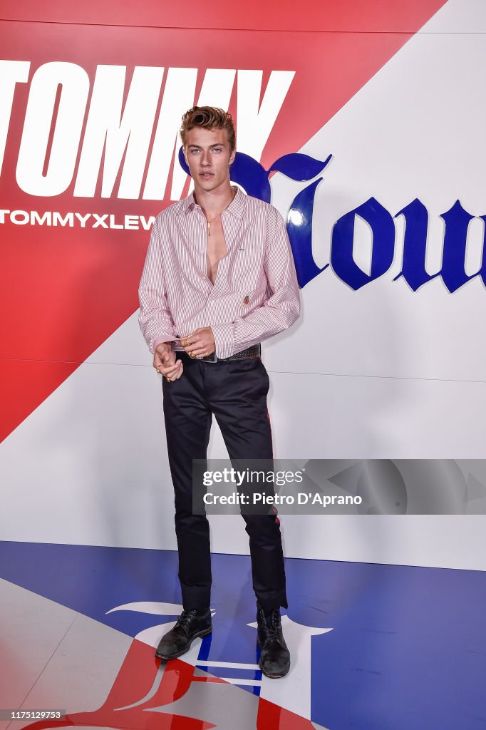 lucky blue smith posing in an open pink and white striped shirt, black dress pants with a red stripe, and ankle boots. he is standing in front of a tommy hilfiger backdrop.