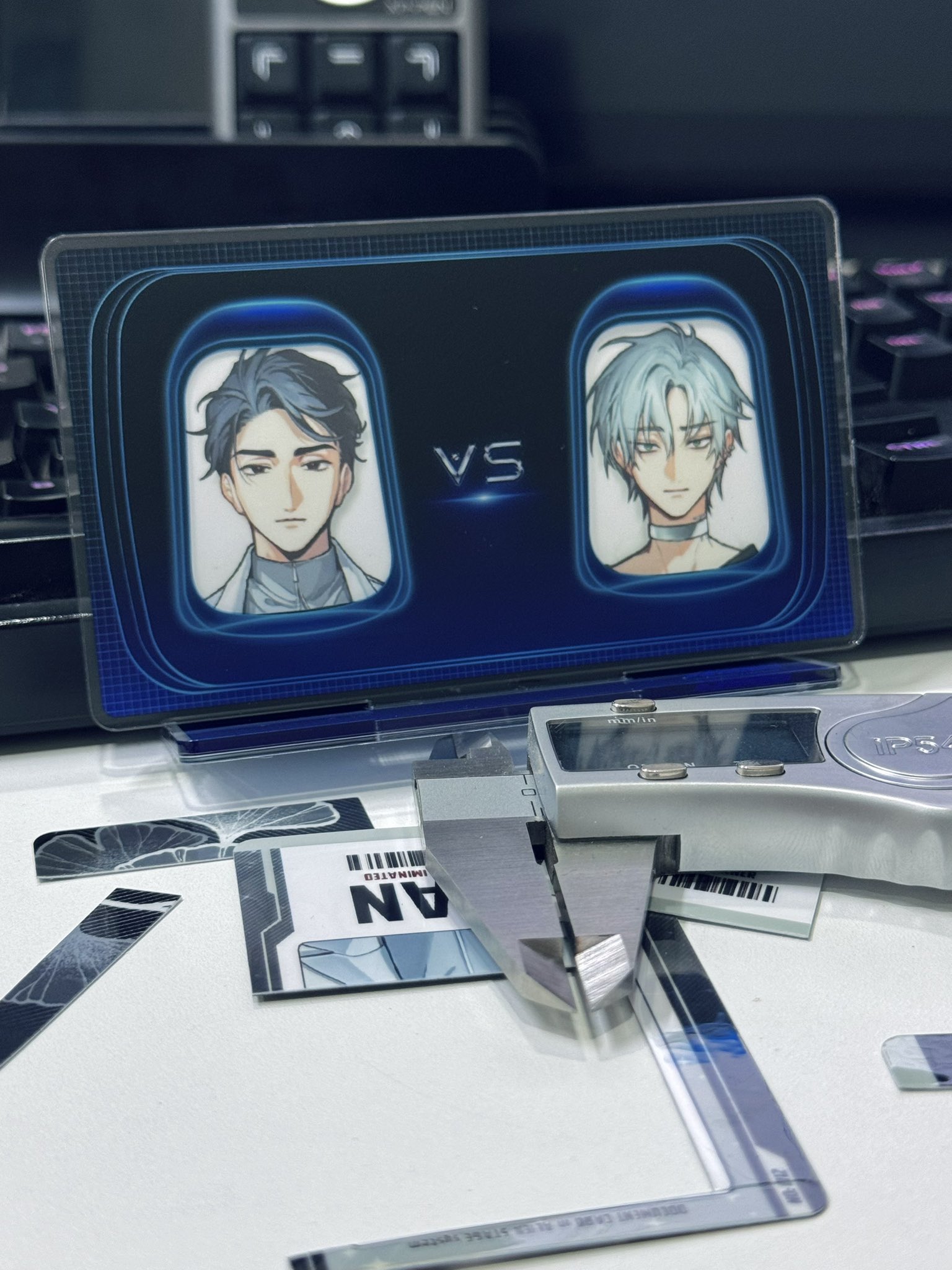 a close up shot of a digital card game card showing two anime boys facing each other with the word vs in the middle. the card is on top of a keyboard and a silver caliper.  the background is blurry.