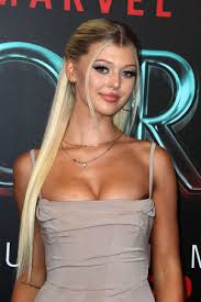 a portrait of loren gray at the thor: love and thunder premiere. she has blonde hair pulled back, wears a light beige dress, and a necklace. the background shows part of the marvel logo and the "thor" title.