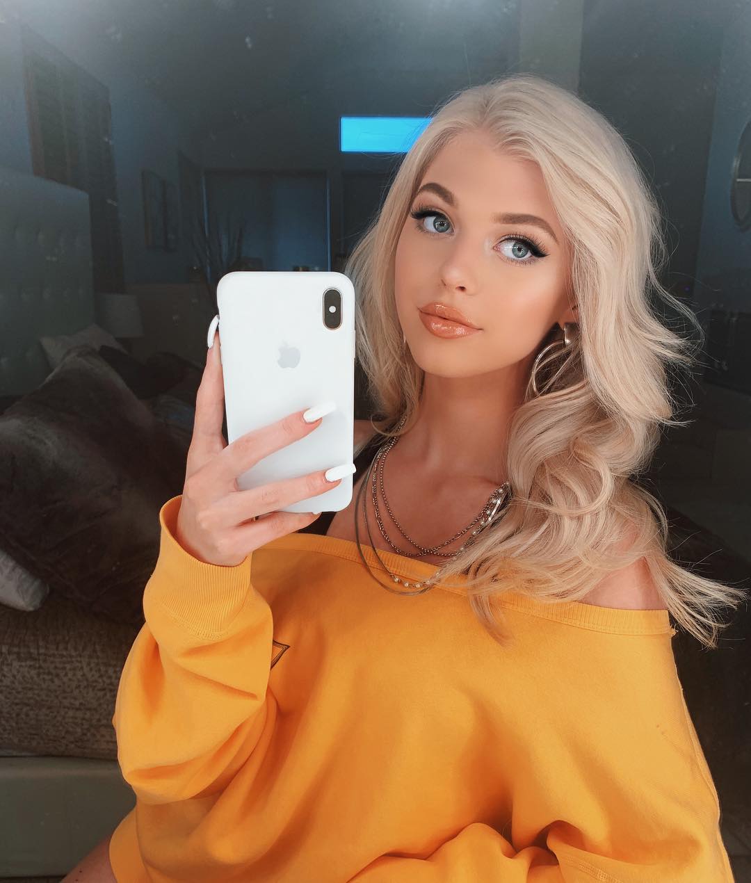 a woman with blonde hair and blue eyes takes a selfie with an iphone. she is wearing an offtheshoulder yellow sweater, a necklace, and earrings. she has long white nails and is wearing makeup. the background appears to be a bedroom with a bed.