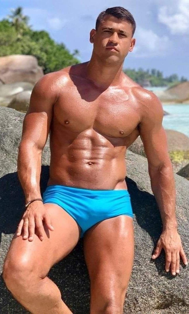 he image shows a muscular, tanned man with a sixpack and toned body relaxing on a rocky beach. he is wearing bright blue swim trunks and has a serious expression on his face. the background is a beautiful sunny beach scene. the man's physique is the main focus of the image.