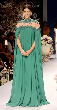 the image shows sonam kapoor, a prominent indian actress, gracefully walking the runway in a stunning teal floorlength gown.  the dress features cape sleeves, a sheer neckline with delicate embroidery, and a flowing, elegant silhouette.  the overall look is sophisticated and luxurious, fitting for a highprofile fashion event.  the background shows a typical fashion show setting, including flowers and other models.