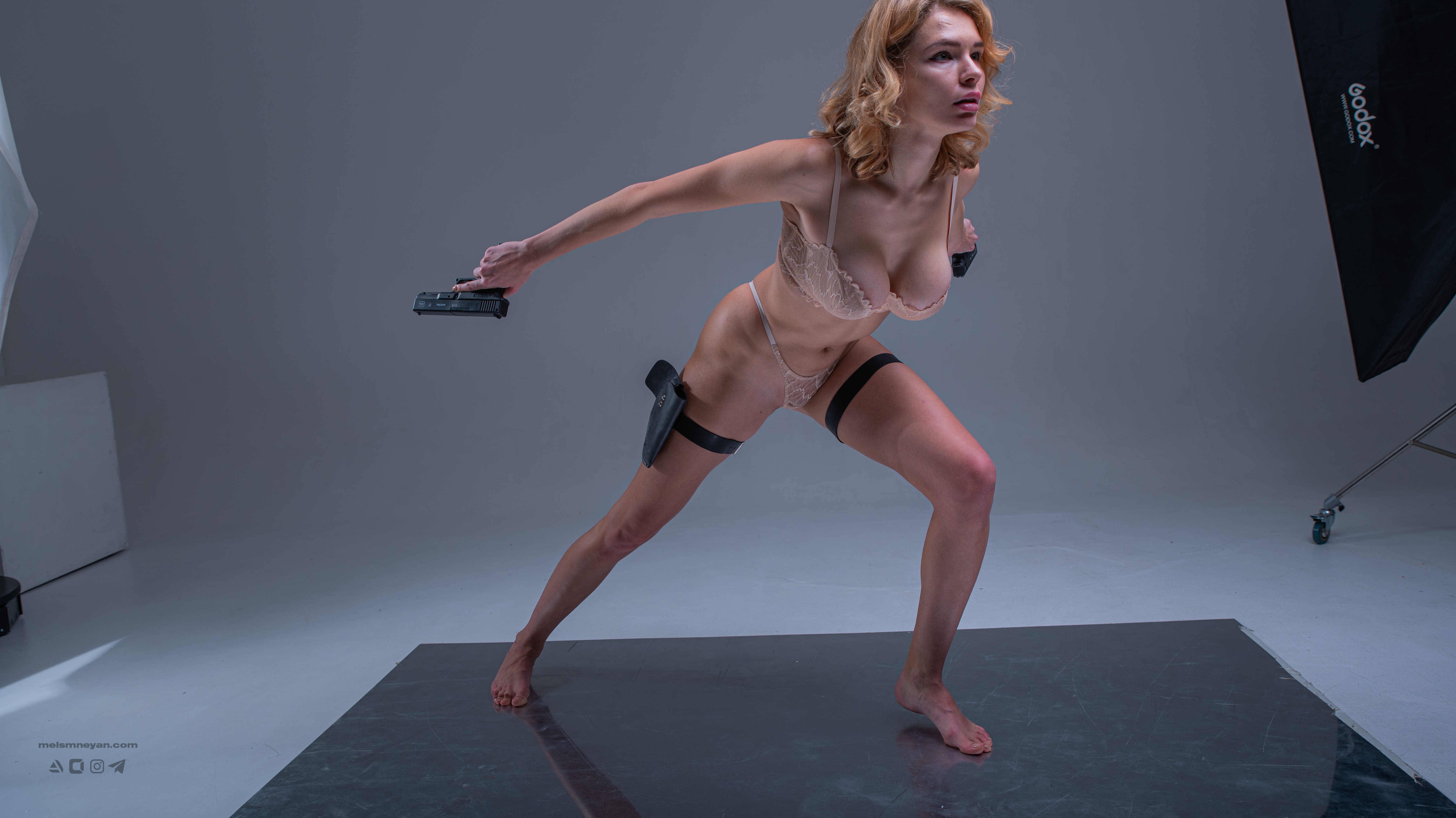 a woman with blonde hair, wearing lingerie and a thigh holster, is posing in a studio. she is holding a gun in one hand and has another gun in her thigh holster. the background is a gray studio setting with some lighting equipment visible. she is barefoot and in an action pose.