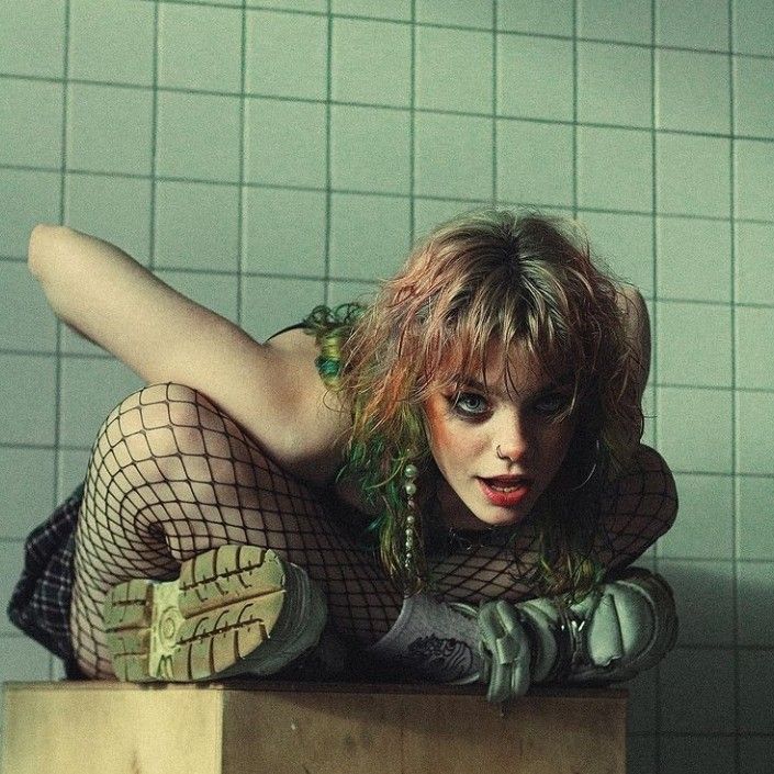 a young woman with green and pink hair is sitting on a box in a bathroom with green tiled walls. she's wearing fishnet tights, a black top, and stylish boots. she's looking directly at the camera with an intense and confident expression. her pose is dramatic and unique, and her overall look is edgy and cool. she's clearly a fashion icon in her own right, and she's not afraid to embrace her individuality.