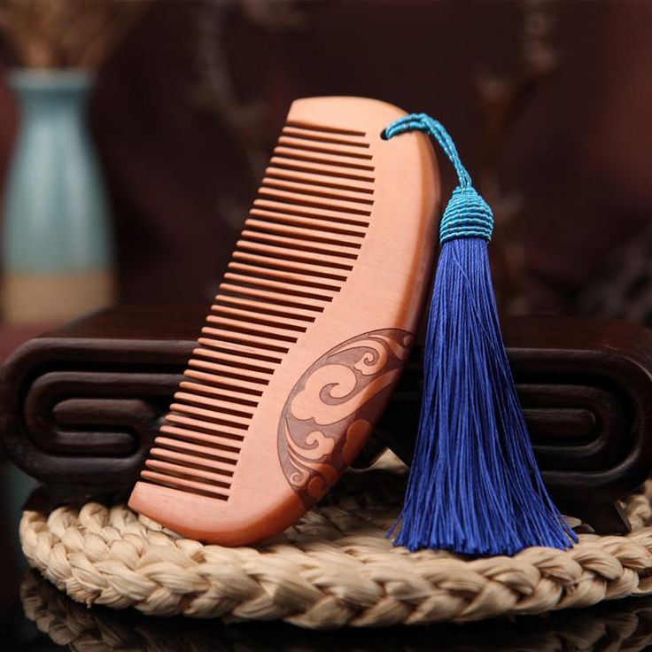 a wooden comb with a blue tassel attached to it is placed on a table. the comb is carved with a design. there is a small vase in the background.