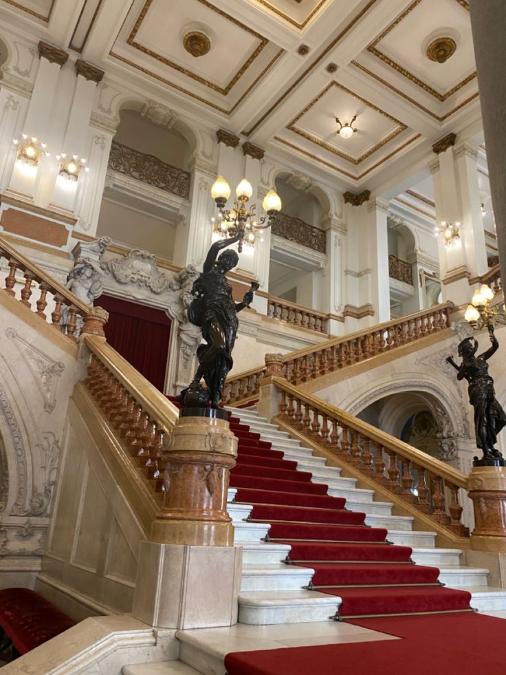 the image shows a grand staircase in a palatial building.  the staircase is made of white marble with a rich red carpet running up its center.  ornate wooden balustrades line the sides.  two classical statues flank the staircase, standing atop marble pedestals.  the ceiling is high and ornately decorated with gold detailing and a chandelier is visible. the overall style is classical and opulent, suggesting a historical building of great significance.  the lighting and composition emphasize the scale and grandeur of the space.