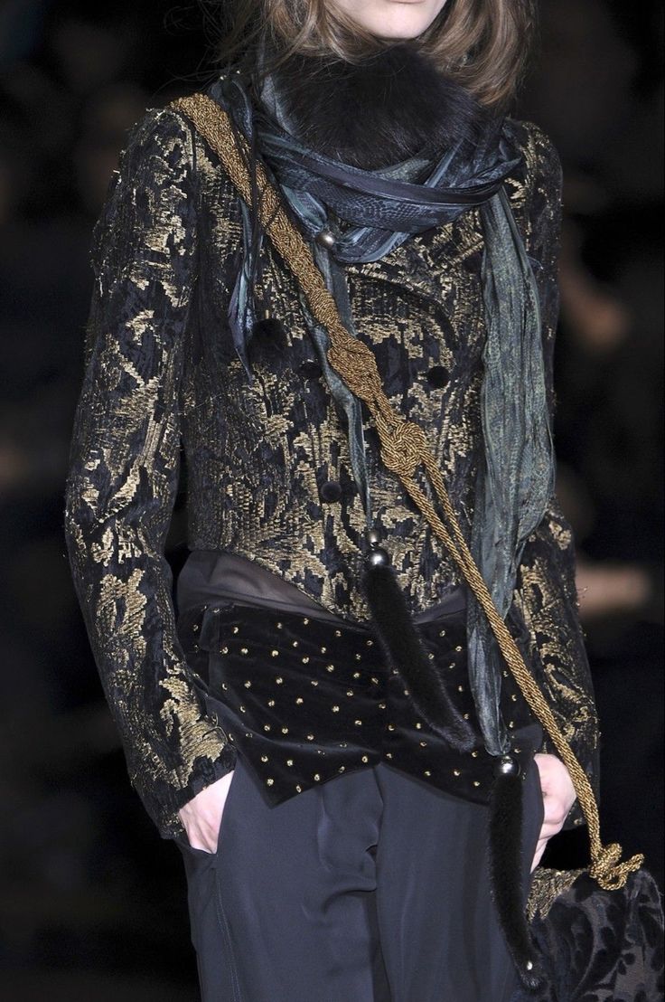 loseup shot of a woman's fashion model on a runway.  she's wearing a dark, brocade jacket with gold detailing, a dark velvet and studded piece underneath, and a long, draped scarf in shades of dark teal and black.  she also carries a dark handbag with goldtoned hardware, and the overall style is sophisticated, elegant, and autumnal. the image focuses on the texture and detail of her clothing and accessories.
