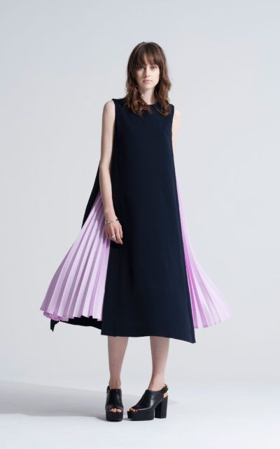 a woman is standing in a studio setting, modeling a stylish sleeveless midi dress. the front of the dress is a solid navy blue, while the sides feature a contrasting lilac pleated skirt that adds a dynamic flair to the design. she is wearing black platform shoes with heels that complement the contemporary look of the dress. the background is a neutral light gray, which helps to emphasize the colors and design of the outfit.