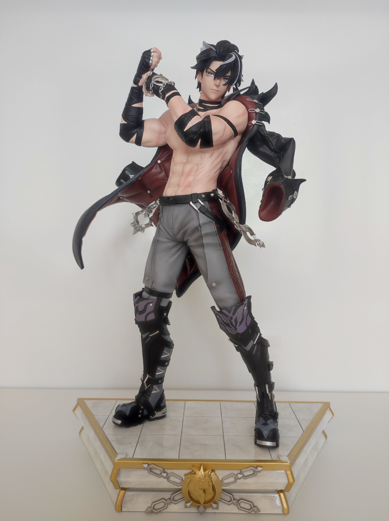 a male anime figurine with black hair standing on a marble base.  he is shirtless, wearing a black leather jacket and grey pants.  he has chains on his waist and has black boots.  the figurine is posing with his right arm in the air and his left fist is clenched.  he is looking directly at the camera.  the base is marblelike and has a gold chain around the edge.  he is a popular character from an anime series.