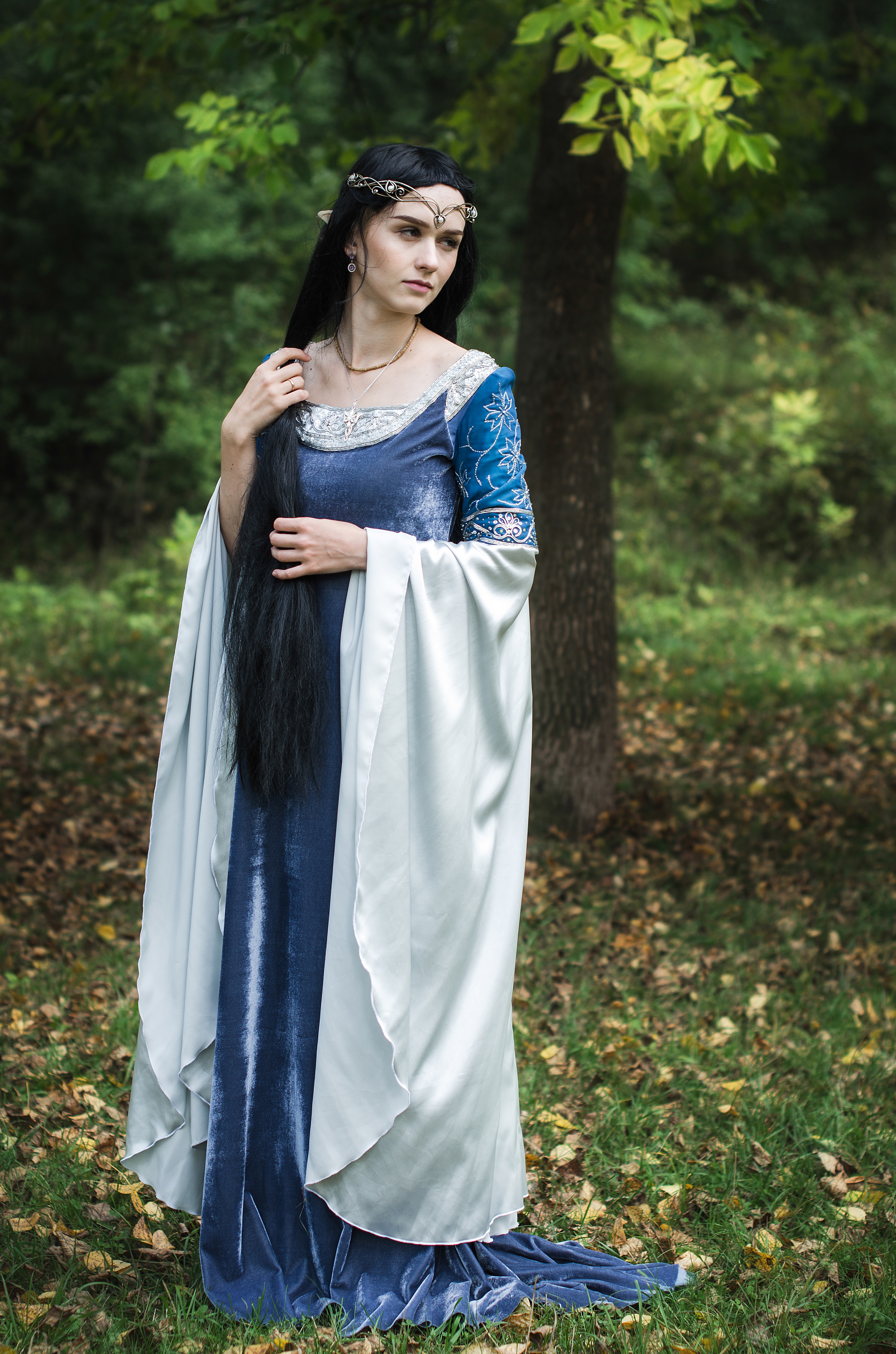 a young woman dressed as an elf stands in a forest. she wears a long, flowing blue velvet gown with a white overdress, and a silver tiara. her long black hair is styled loosely. she looks beautiful and graceful, and her pose suggests a sense of quiet contemplation. the setting is autumnal, with fallen leaves scattered on the ground. the overall aesthetic is one of fantasy and fairytale.