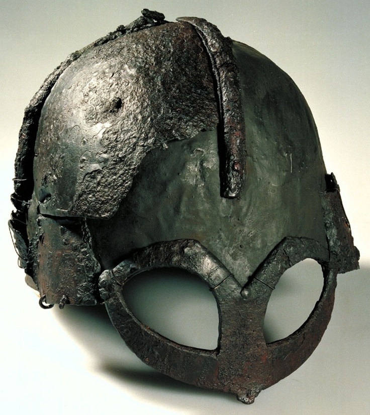the image shows an iron viking helmet with a spectacle guard that is rusted and has a patch on the top. the helmet is lit from the front with studio lighting and is shot from a slightly elevated side perspective.