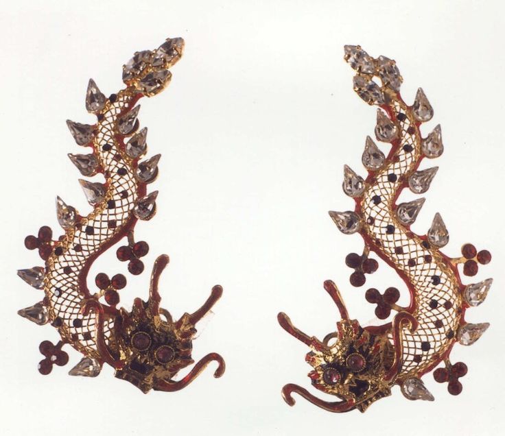 two ornate, dragonshaped earrings are displayed on a white background. they feature a goldtone filigree design with red accents, and are embellished with clear gemstones and crystals. the earrings have a dragonlike head with curled features and several floral or clover shaped embellishments.