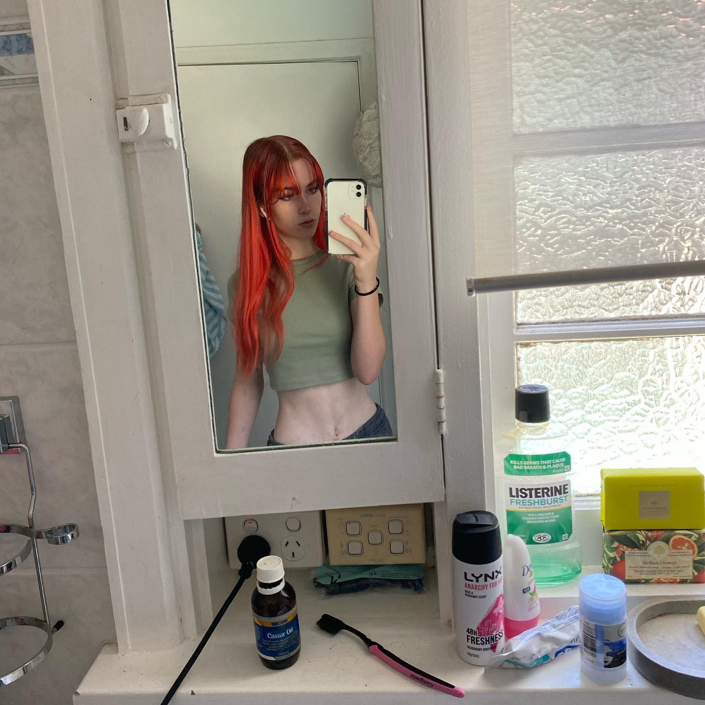 a young woman with long, bright red hair and a casual outfit (green crop top and grey sweatpants) takes a mirror selfie in a brightly lit bathroom. the photo shows the reflection of part of her bathroom, including a window, a medicine cabinet, a toothbrush holder, and various toiletries on the counter.
