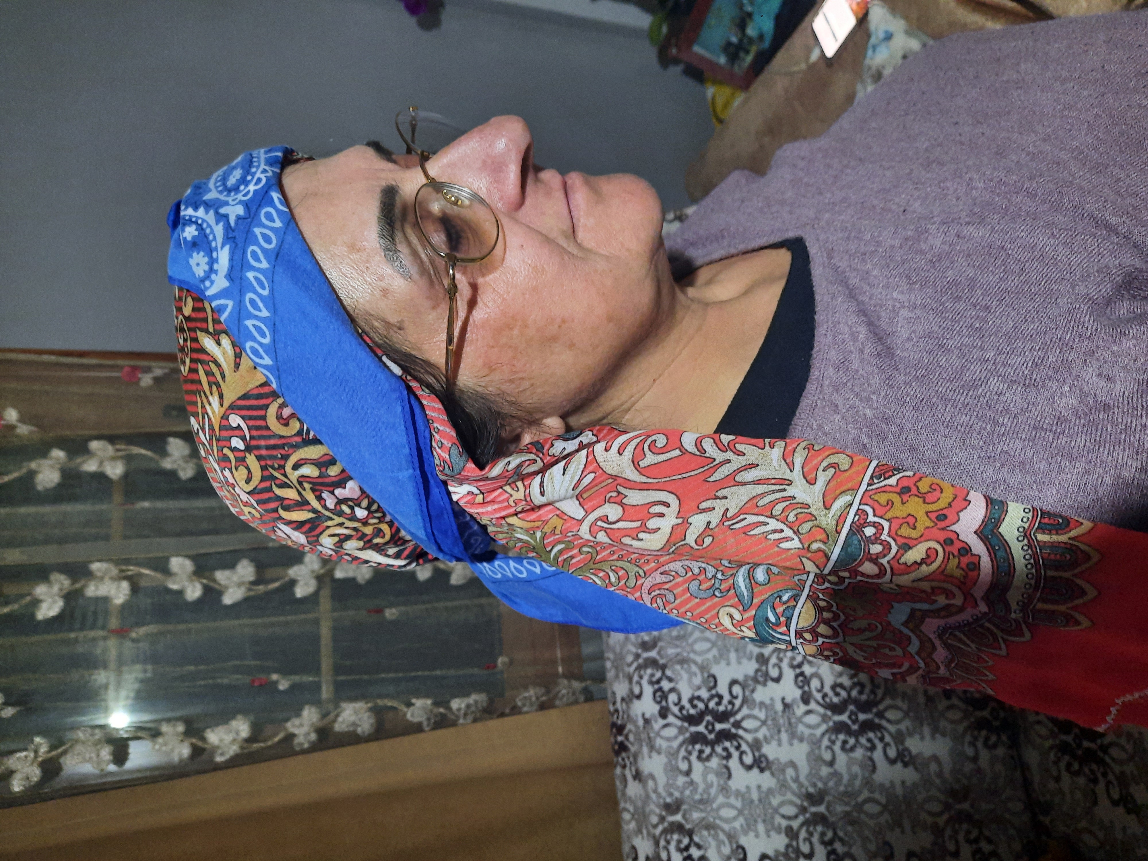 a middleaged woman wearing a colorful patterned headscarf and glasses is sitting indoors. the headscarf is layered with different patterns and includes a blue fabric section. she is wearing a purple shirt. behind her, there are patterned curtains by a window and a couch. the photo is a profile shot of the woman.