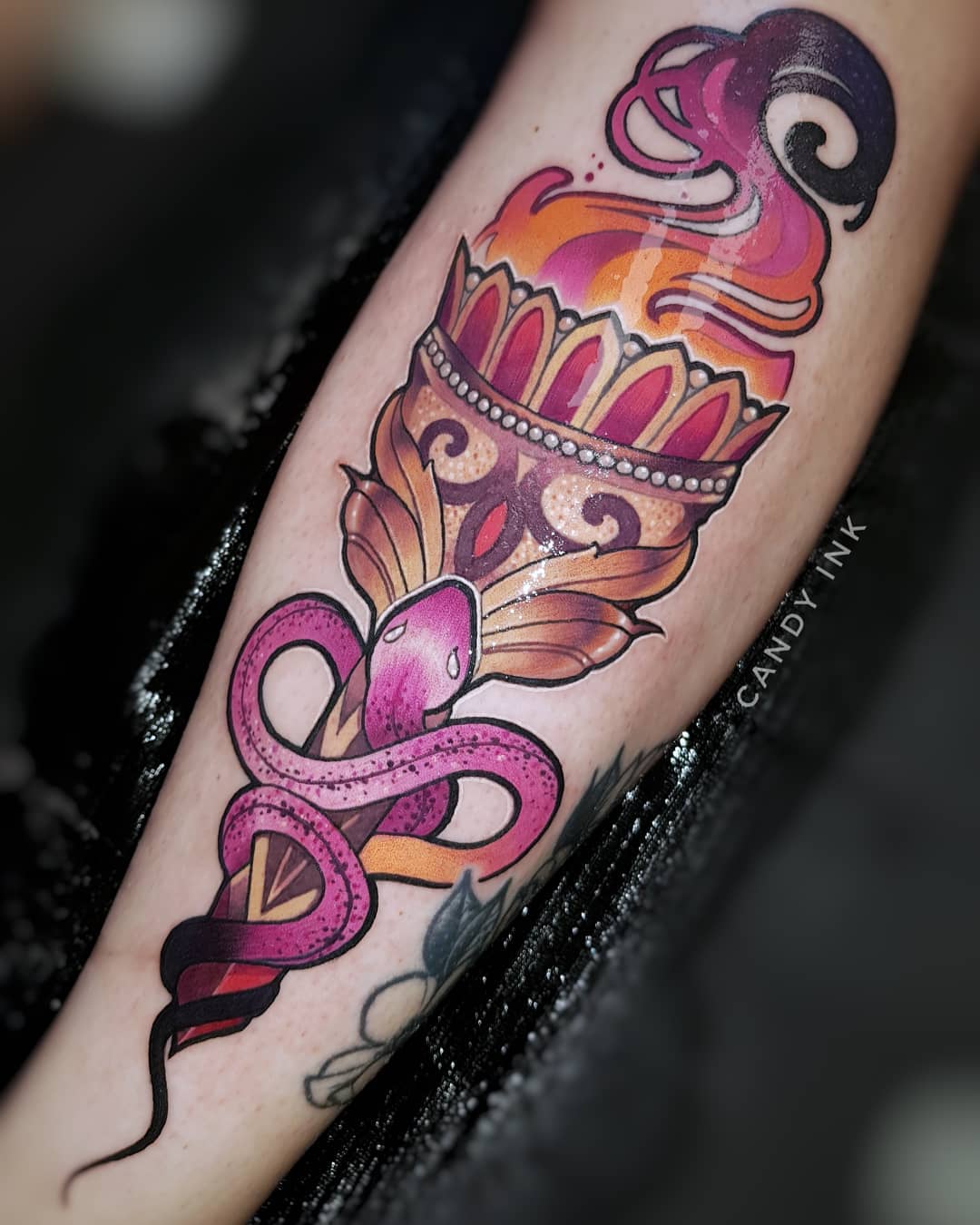 the image showcases a vibrant neotraditional tattoo of a burning torch entwined with a snake. rendered in a rich color palette, the tattoo features a golden torch with intricate detailing, engulfed in stylized flames of purple, orange, and yellow. a pink snake with black dotted scales wraps around the torch's base, its tail ending near the viewer. the torch is adorned with jewellike elements, adding to its visual appeal. the tattoo sits on the inner forearm of a person, the artist's mark "candy ink" subtly placed below the design.